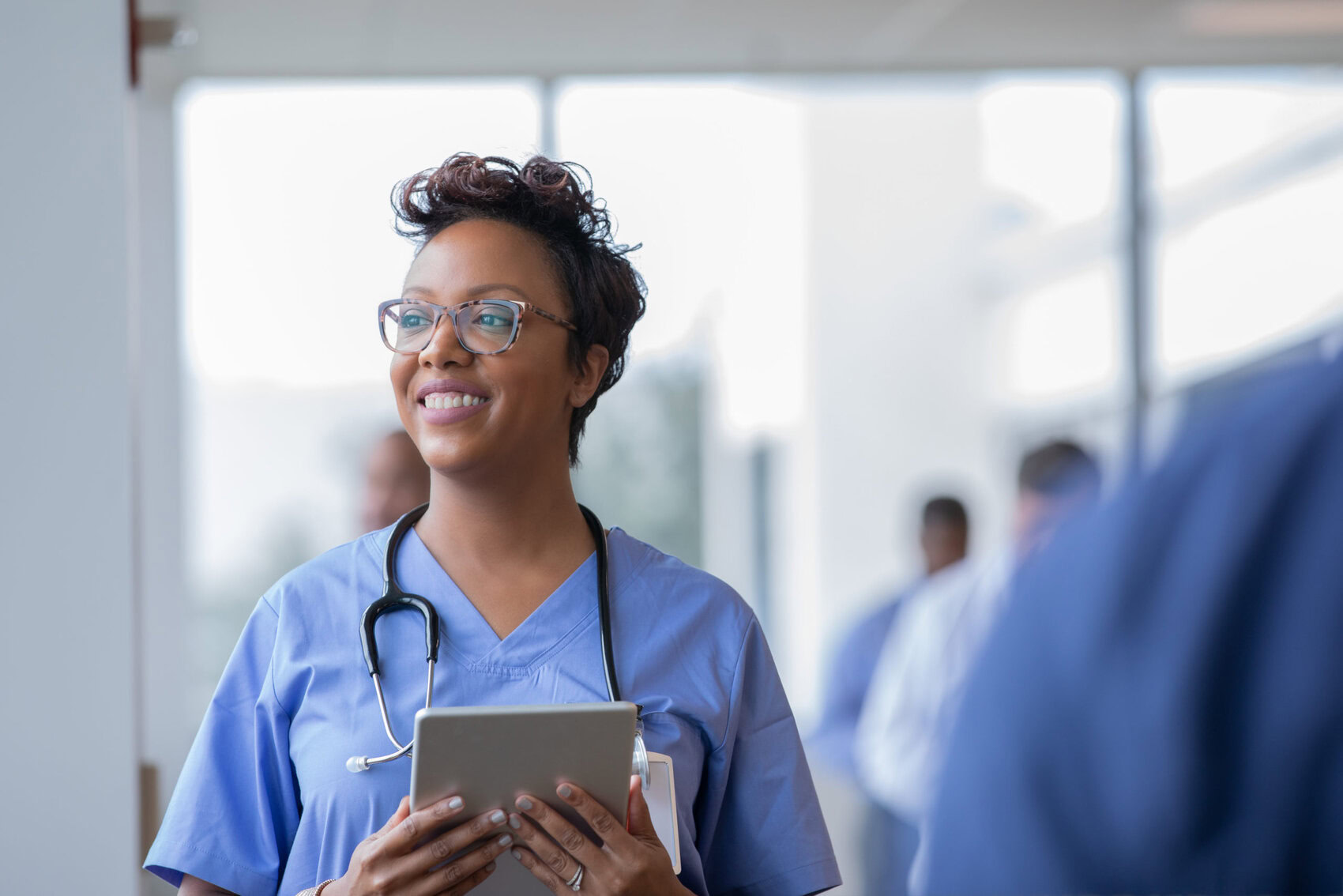 Female nurse or doctor smiles while staring out window in hospital hallway and holding digital tablet with electronic patient file Female nurse or doctor smiles while staring out window in hospital hallway and holding digital tablet with electronic patient file