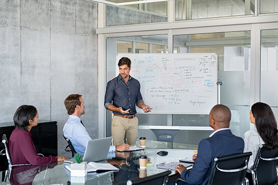 Man in front of white board presenting to four colleagues seated at table