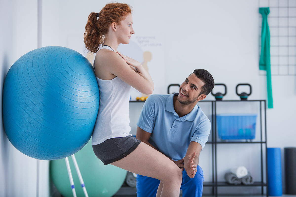 Woman leaning on exercise ball while physical therapist kneels to help