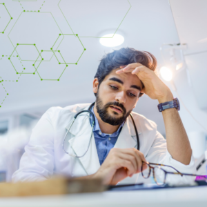 Medical resident in scrubs and white coat at desk, appearing overwhelmed with head in hand.